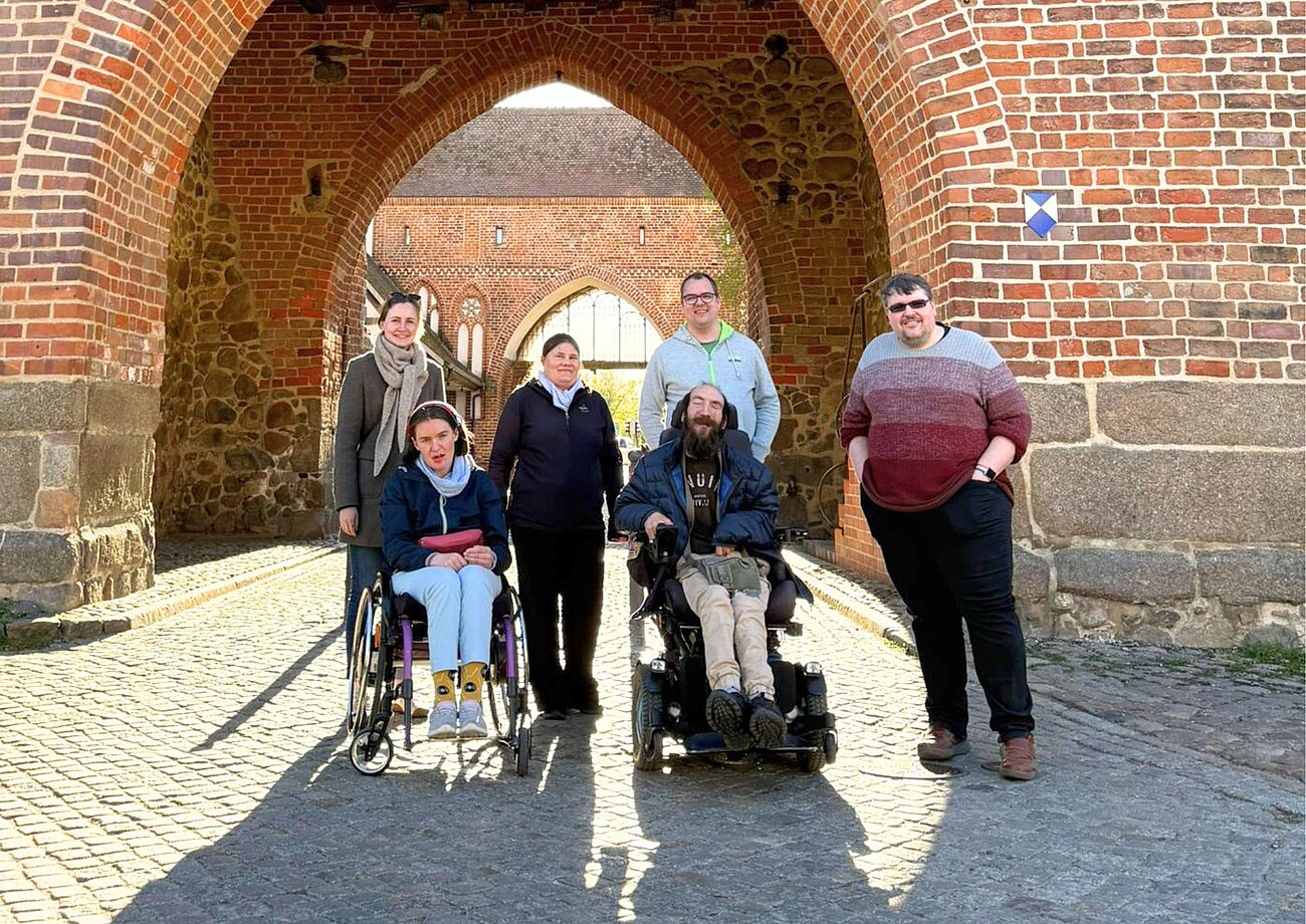 6 Personen stehen vor einem Tor. 2 Personen sitzen in einem Rollstuhl.